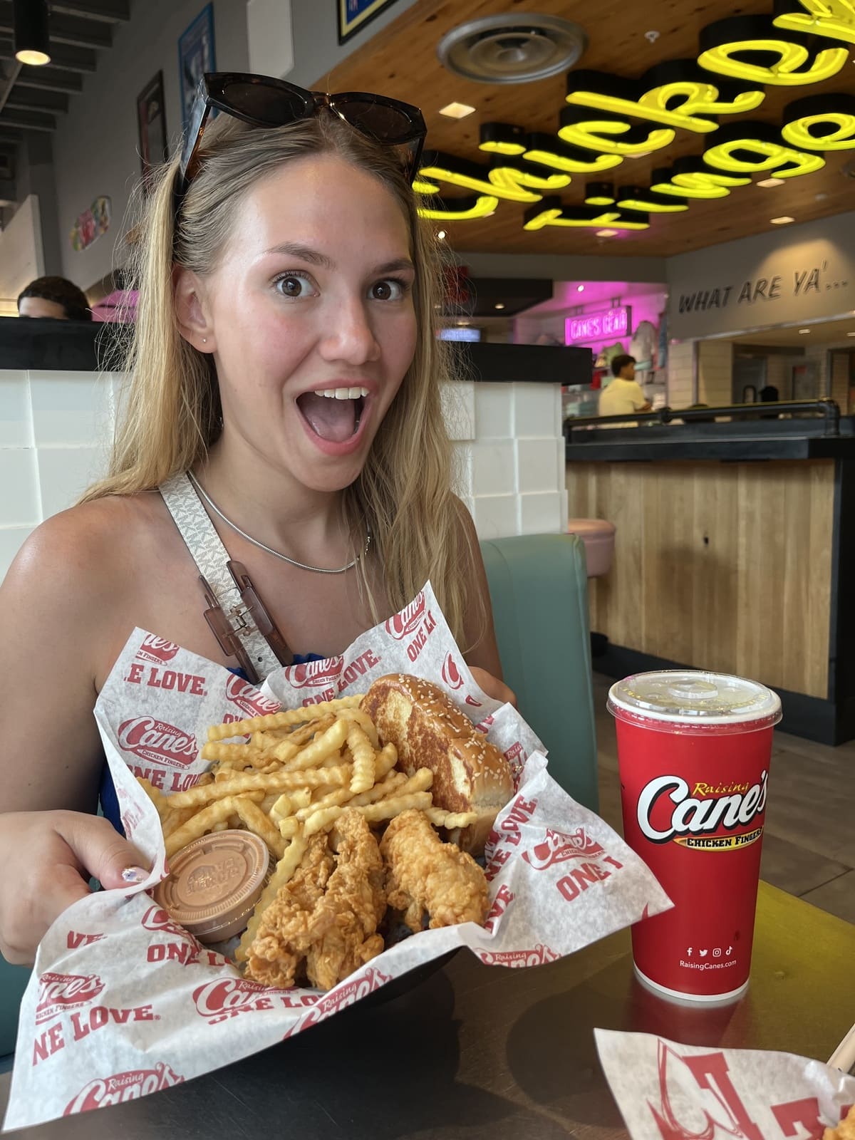 Smiling woman holding a basket of Raising Cane’s chicken fingers, crinkle fries, dipping sauce, and a sesame seed bun with a Raising Cane’s drink cup on the table.