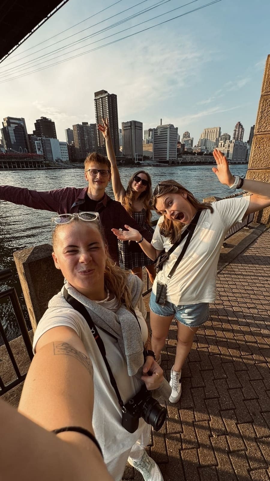 Group of four young friends posing energetically for a selfie by a riverside with city skyscrapers in the background during daylight.