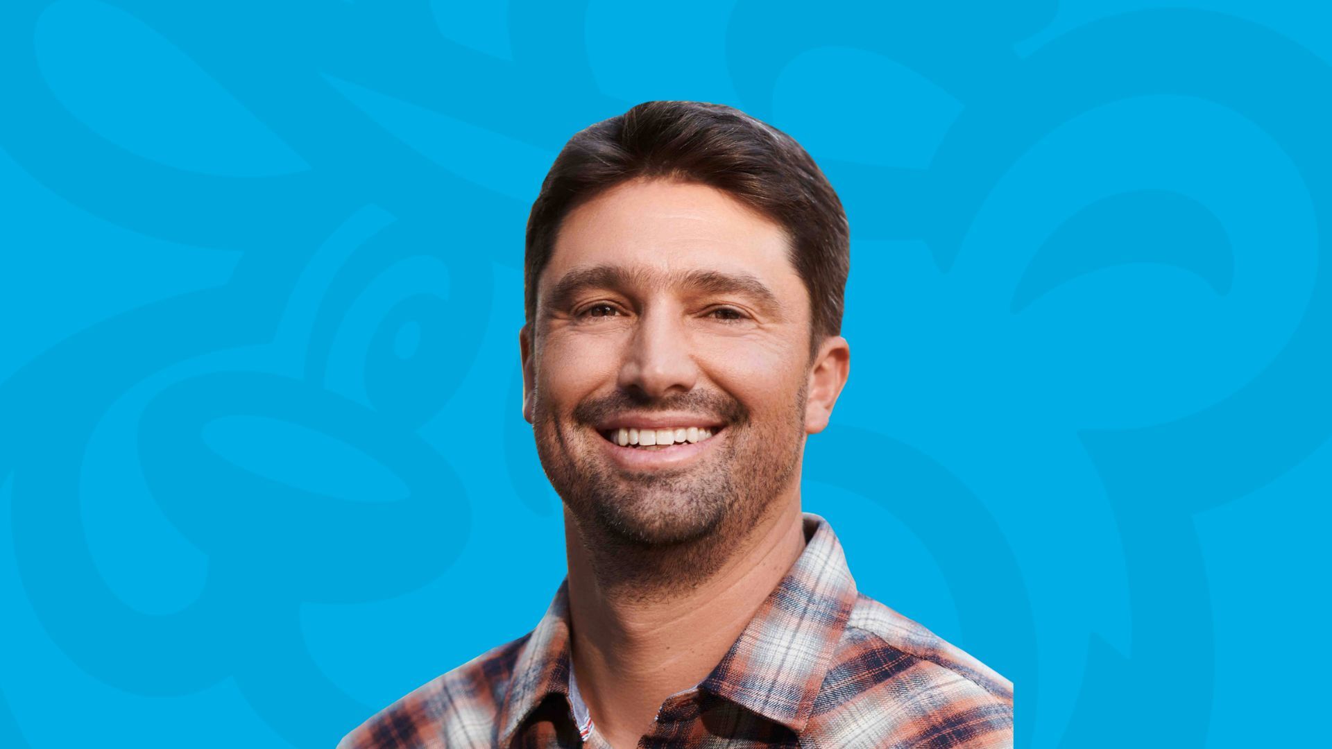 Smiling man with short dark hair and a beard wearing a plaid shirt against a blue background.