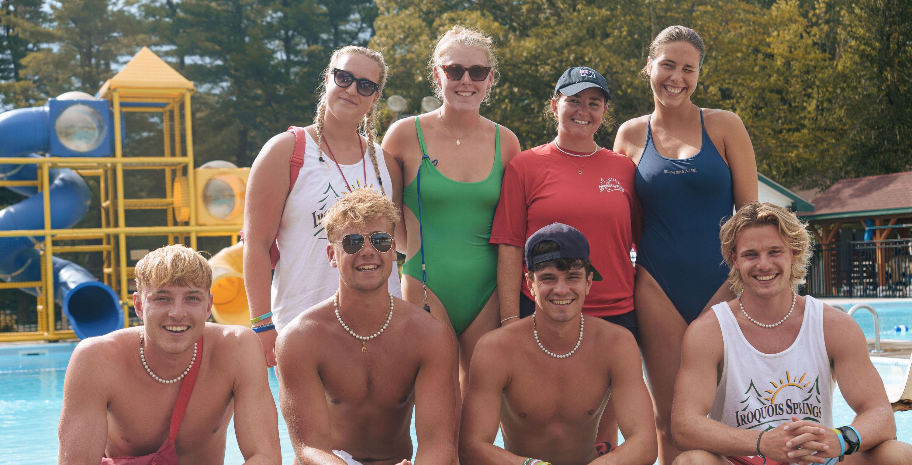 Group of seven young lifeguards smiling by a pool with a yellow and blue water slide in the background.