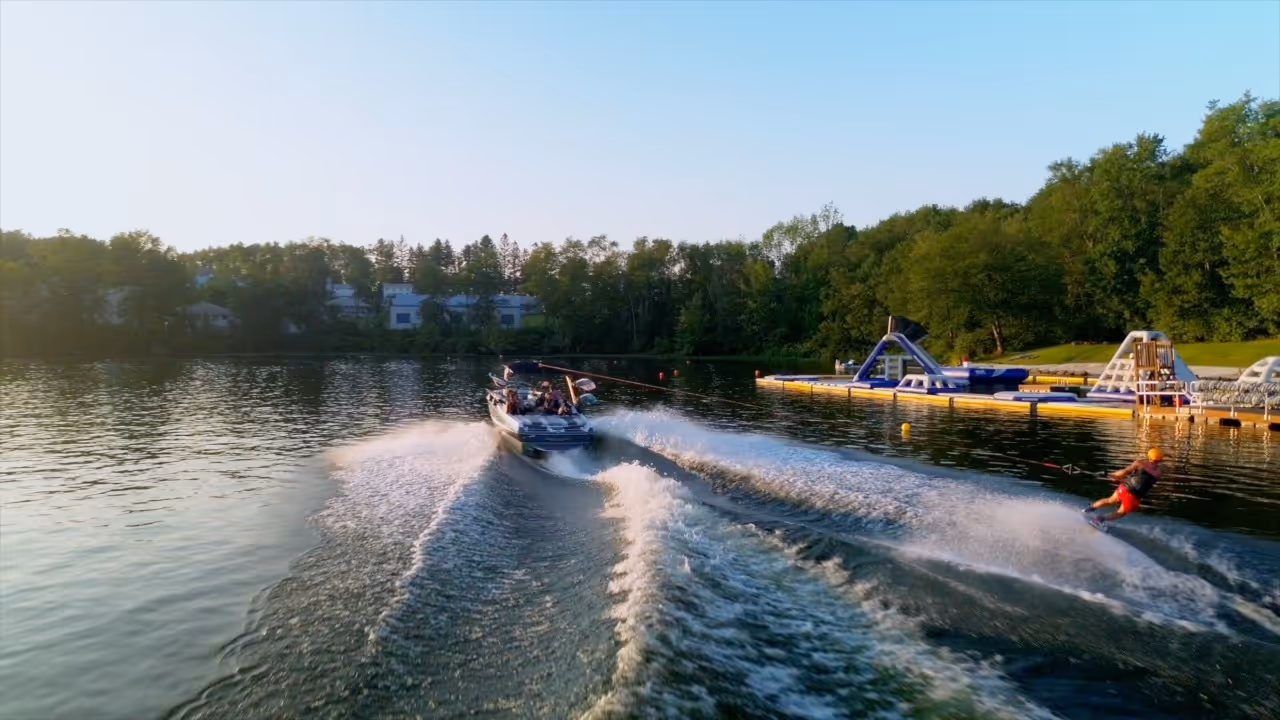Person wakeboarding on a lake, being pulled by a boat with passengers, near an inflatable water park and surrounded by trees.
