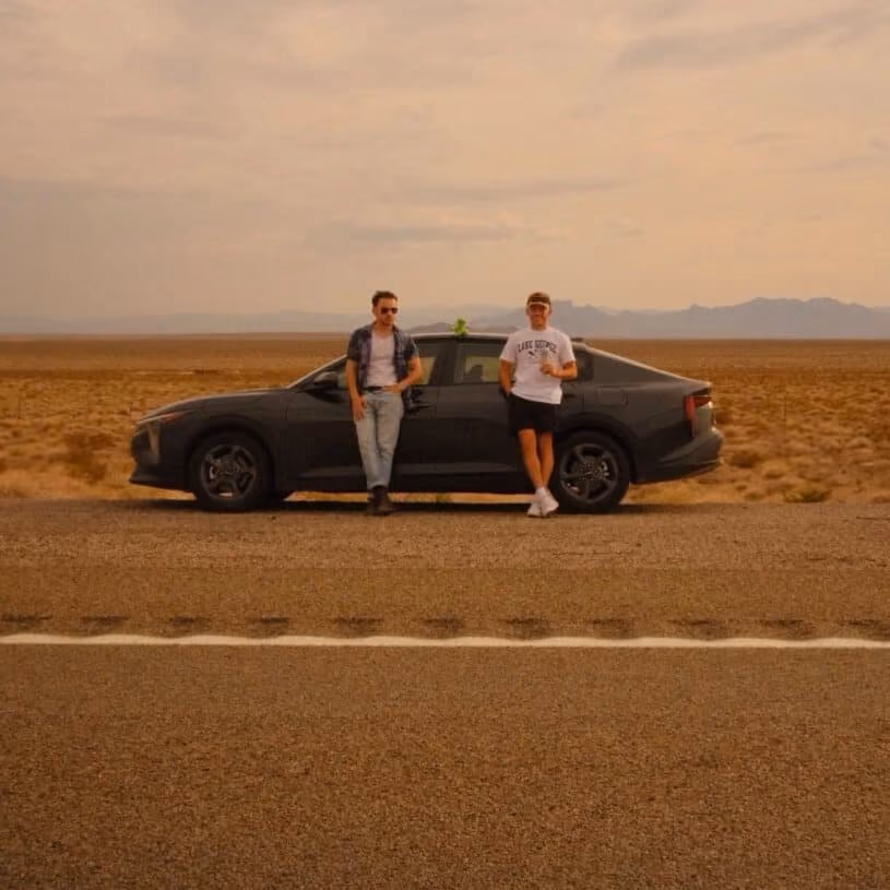 Two men standing beside a black car parked on the roadside in a desert landscape with mountains in the distance.