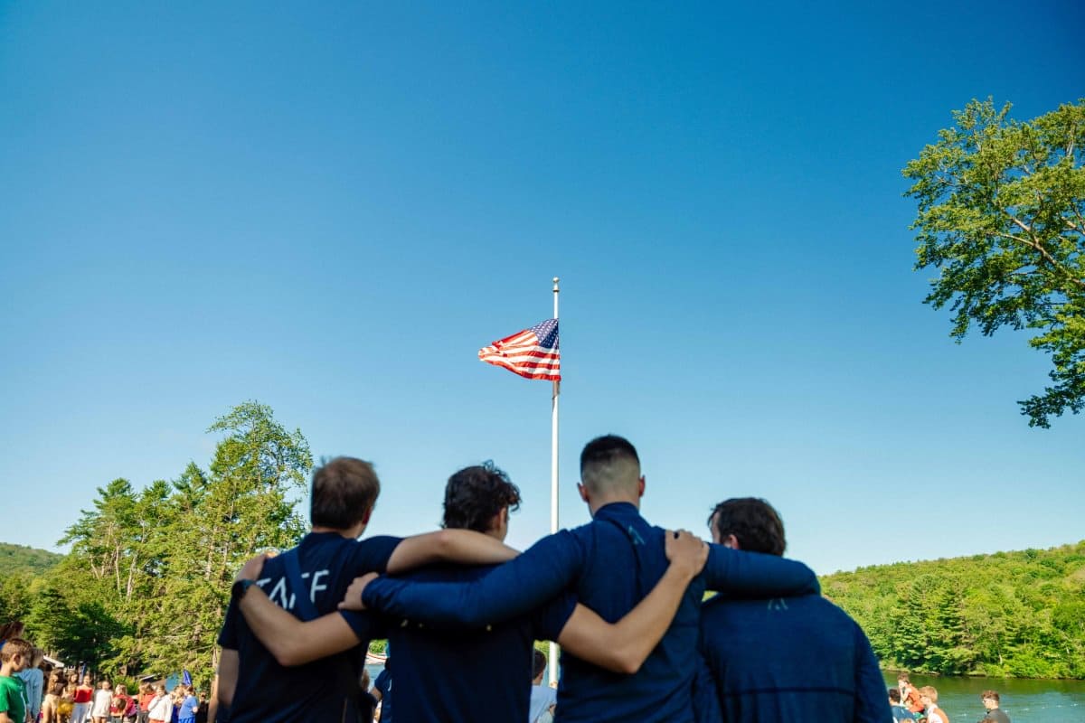 Four people with arms around each other facing an American flag on a sunny day by a lake.
