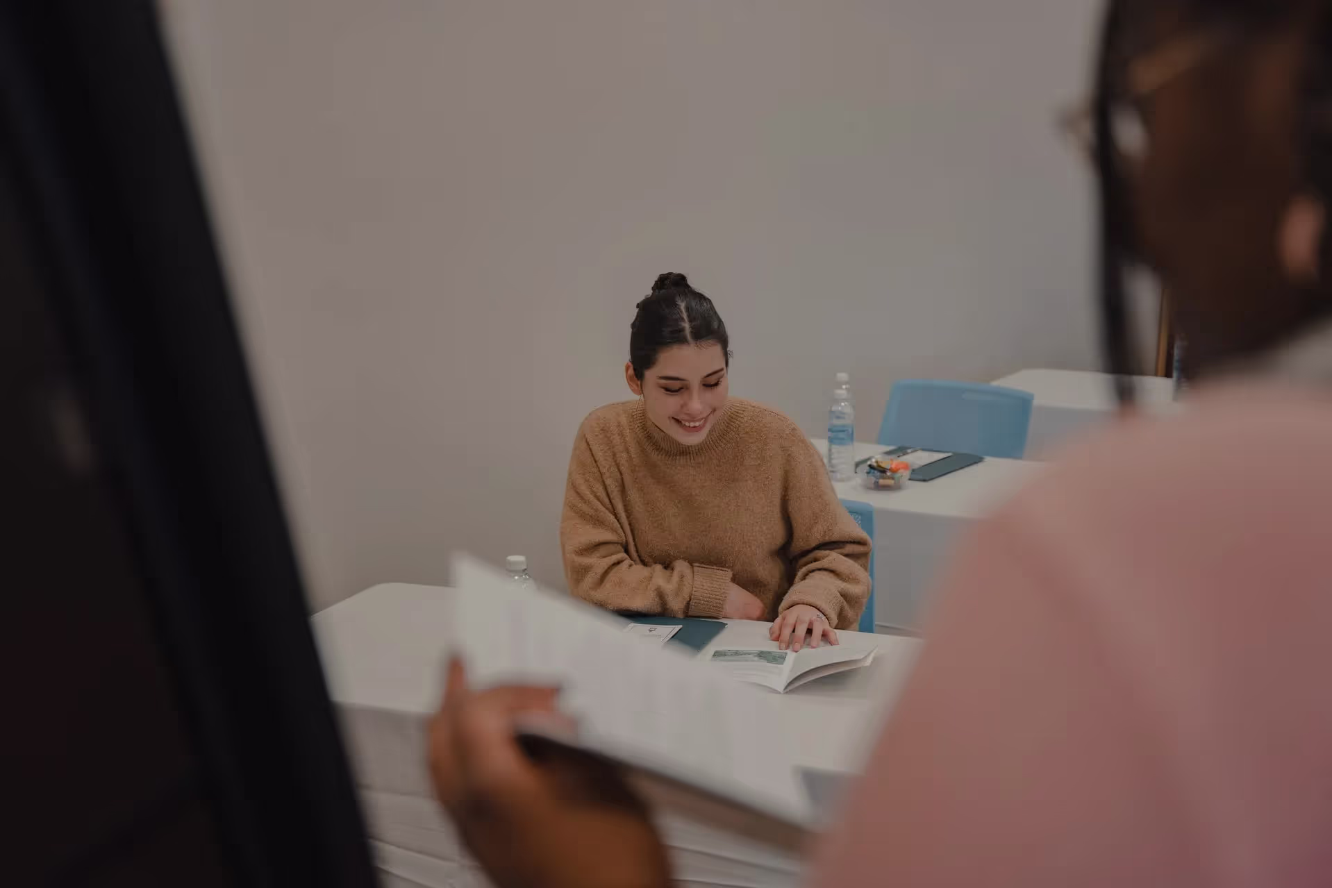sitting in a room, writing notes, while a woman in the foreground is speaking