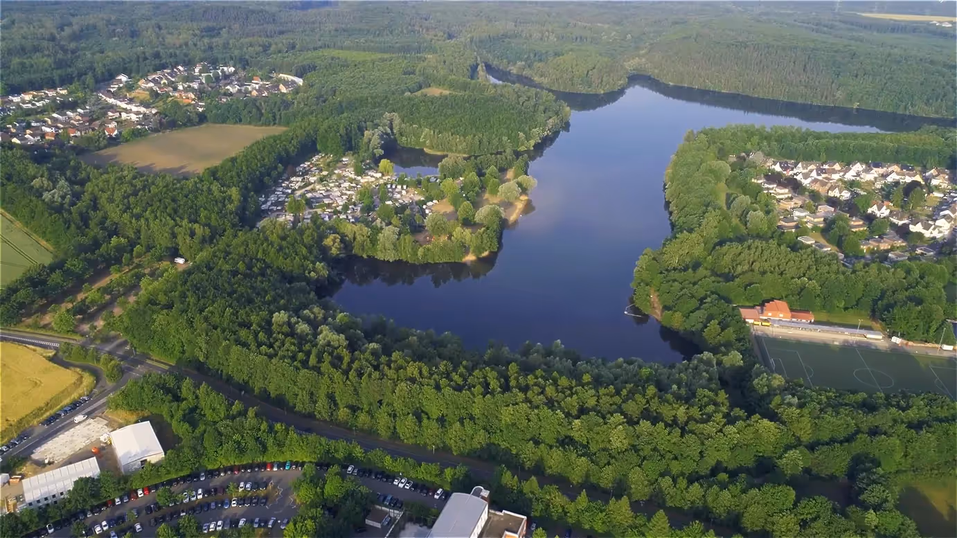 Luftaufnahme eines großen Sees umgeben von dichtem Wald, Wohnsiedlungen und Sportplatz bei sonnigem Wetter.