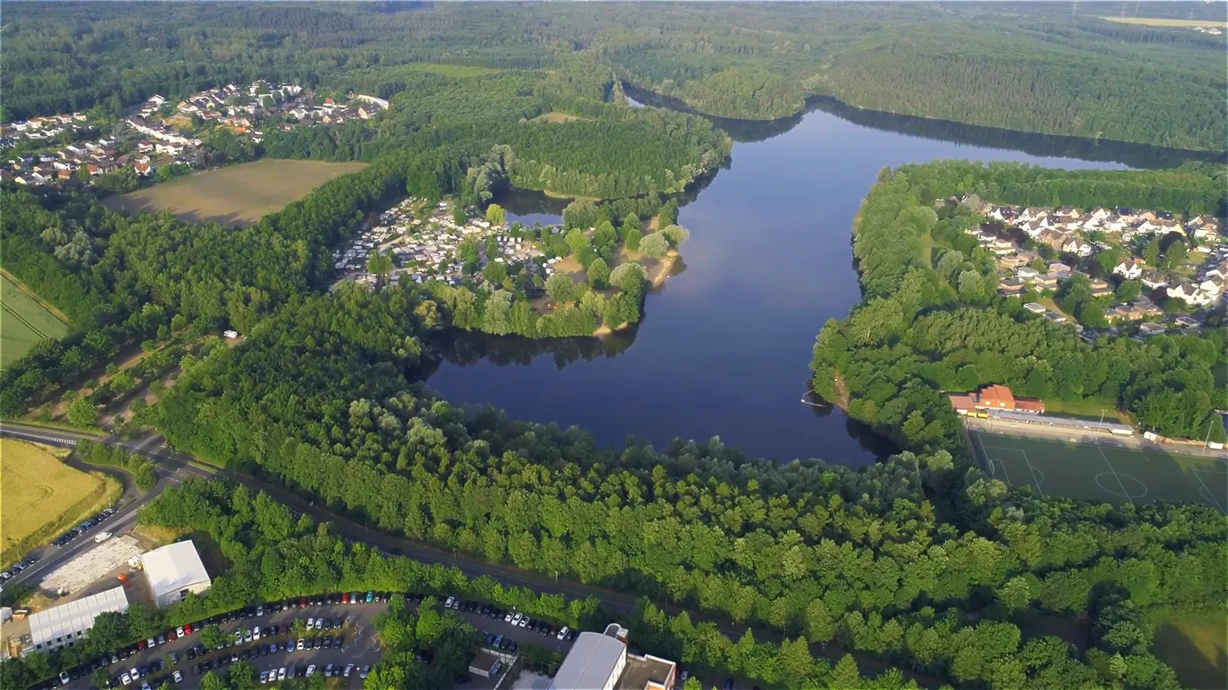 Luftaufnahme eines großen Sees umgeben von dichtem Wald, Wohnsiedlungen und Sportplatz bei sonnigem Wetter.