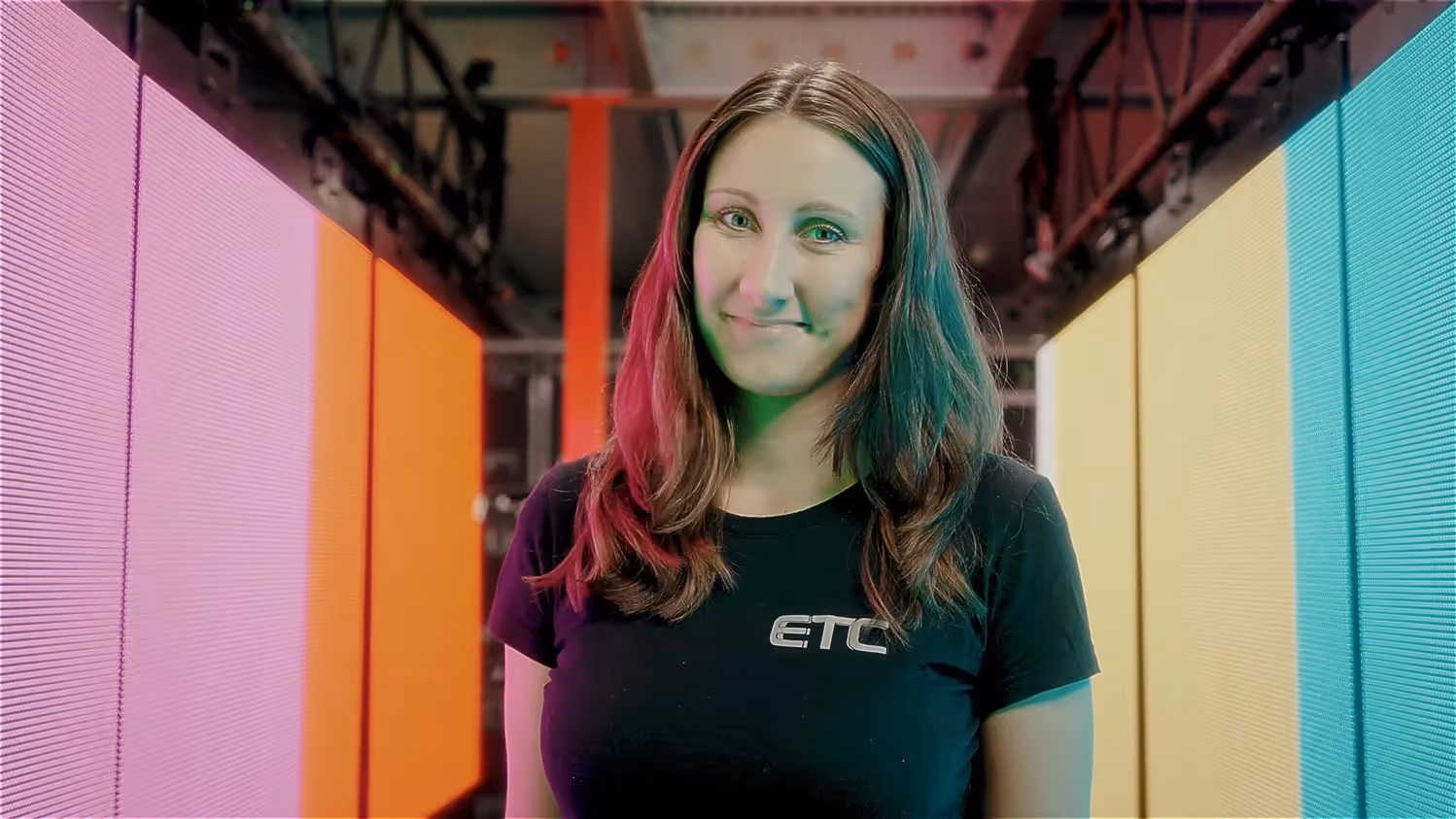 Junge Frau im schwarzen T-Shirt mit ETC-Logo steht zwischen bunten LED-Wänden in Pink, Orange, Gelb und Blau, die ein modernes Lichtambiente erzeugen.