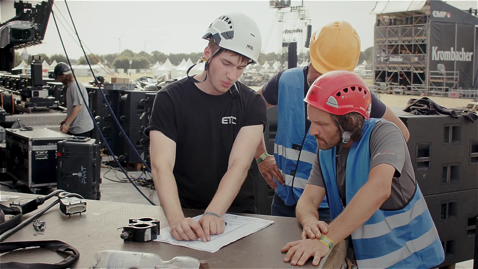 Drei Techniker mit Helmen und Warnwesten besprechen einen Bauplan auf einem Tisch bei einer Festival-Bühne. Der Mann links zeigt mit dem Finger auf den Plan, während die anderen konzentriert zuhören. Im Hintergrund Bühnentechnik, Kabel und Traversen.