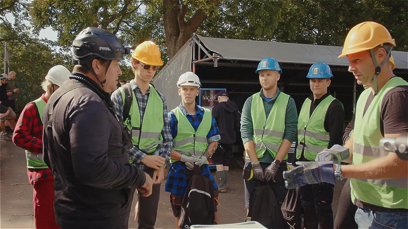 Eine Gruppe von Veranstaltungstechnikern mit Schutzhelmen und Warnwesten bei einer Sicherheits- oder Einsatzbesprechung im Freien. Im Hintergrund sind Zelte und Bäume zu sehen.