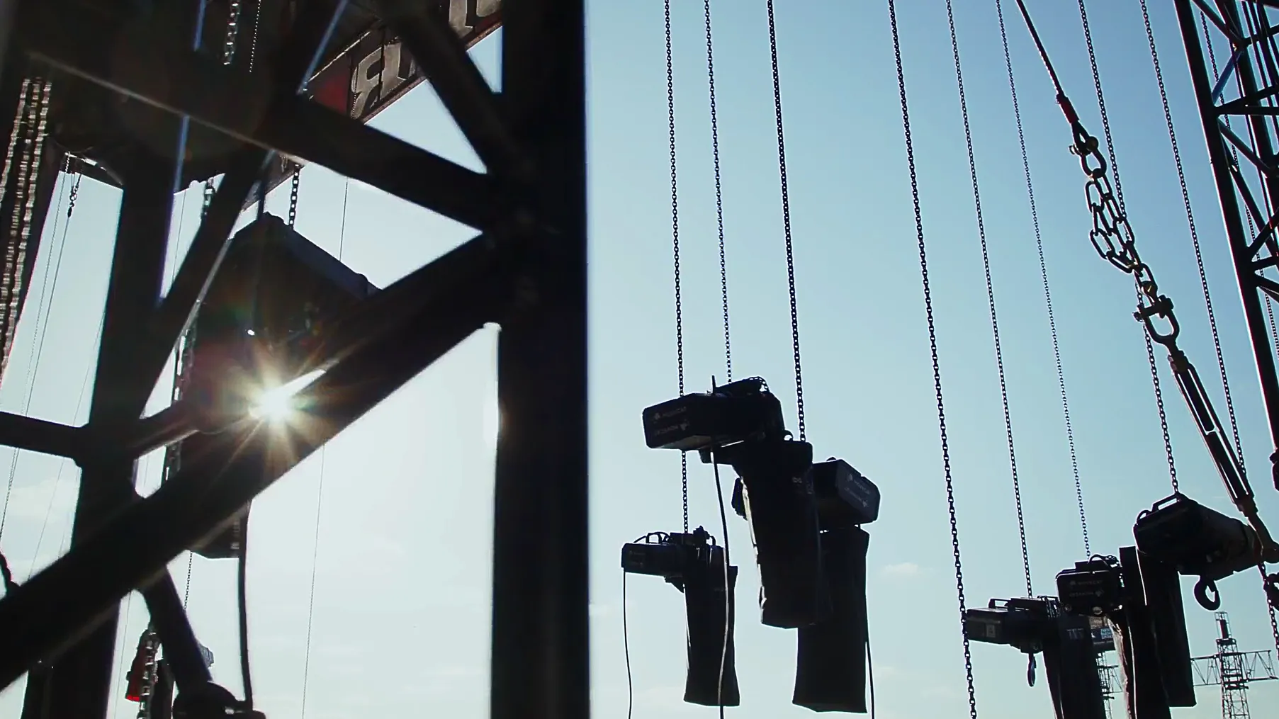 ChatGPT:  Alt-Text-Vorschlag: „Blick auf eine Bühnenkonstruktion mit schwarzen Traversen, Seilen und hängenden Scheinwerfern, die an Ketten befestigt sind. Im Hintergrund scheint die Sonne durch die Metallstreben.
