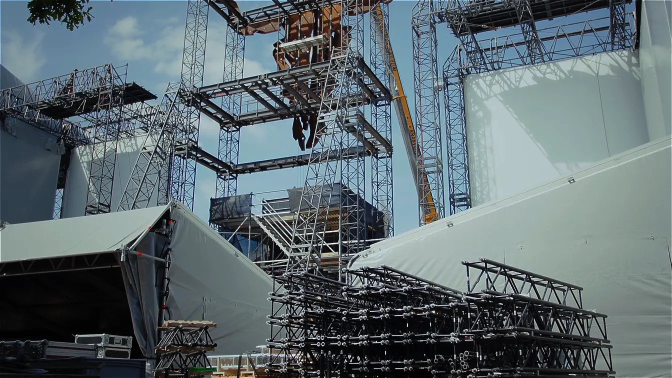 Großaufbau einer Festival-Bühne mit Stahltraversen, Gerüsten und Kränen. Im Vordergrund liegen gestapelte schwarze Traversenteile, im Hintergrund sind Treppen, Podeste und weiße Abdeckplanen zu sehen.