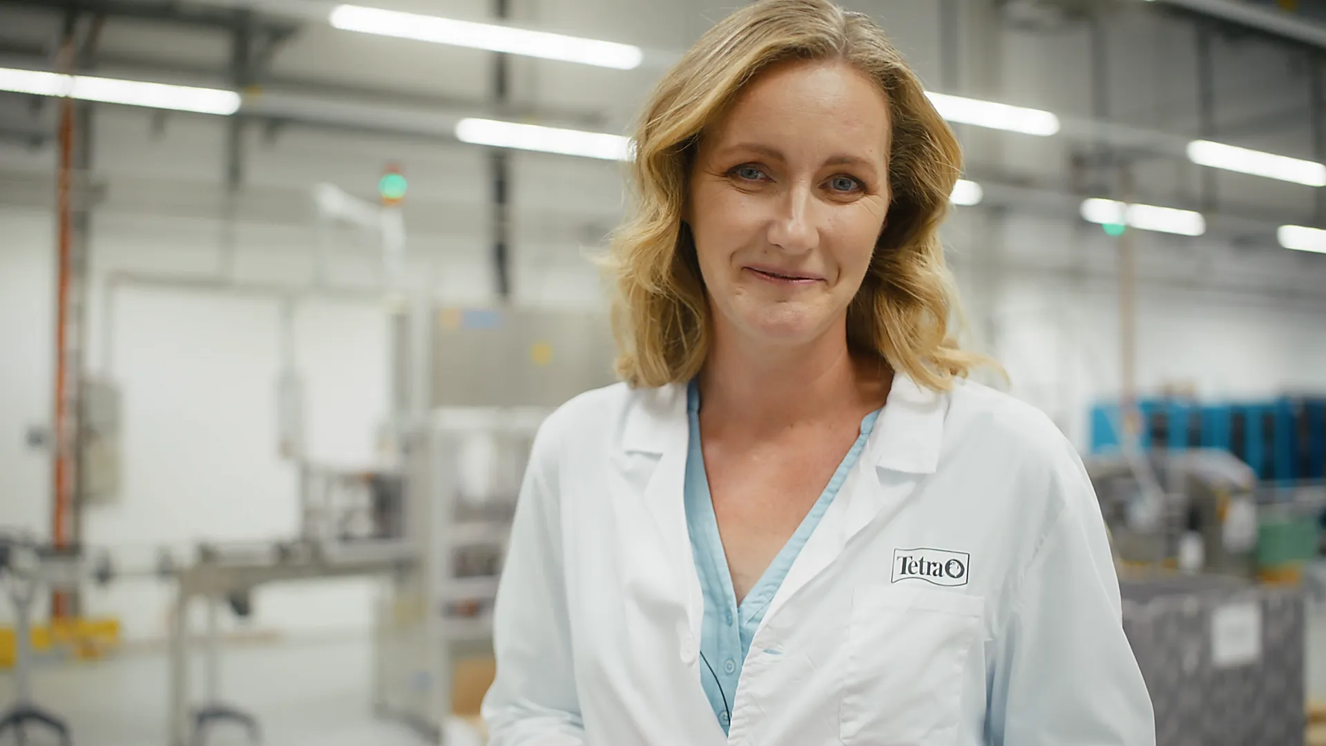 Porträt einer Wissenschaftlerin in weißem Laborkittel mit Tetra-Logo, stehend in moderner Produktionshalle, Realfilm, natürliche Farben.
