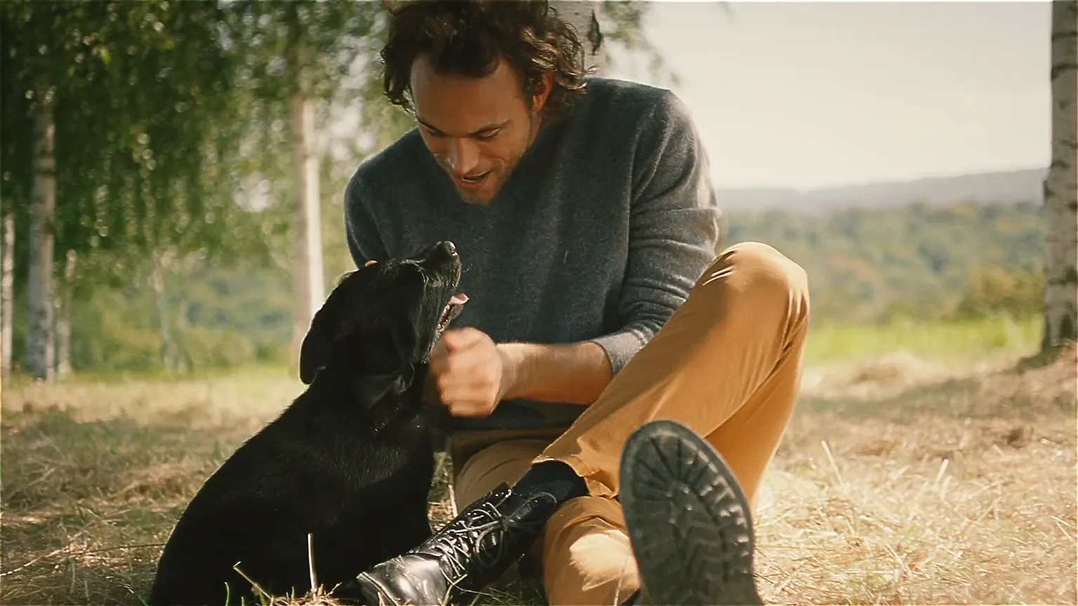 Person sitzt auf einer Wiese und spielt fröhlich mit einem schwarzen Hund unter blauem Himmel mit Bäumen im Hintergrund.