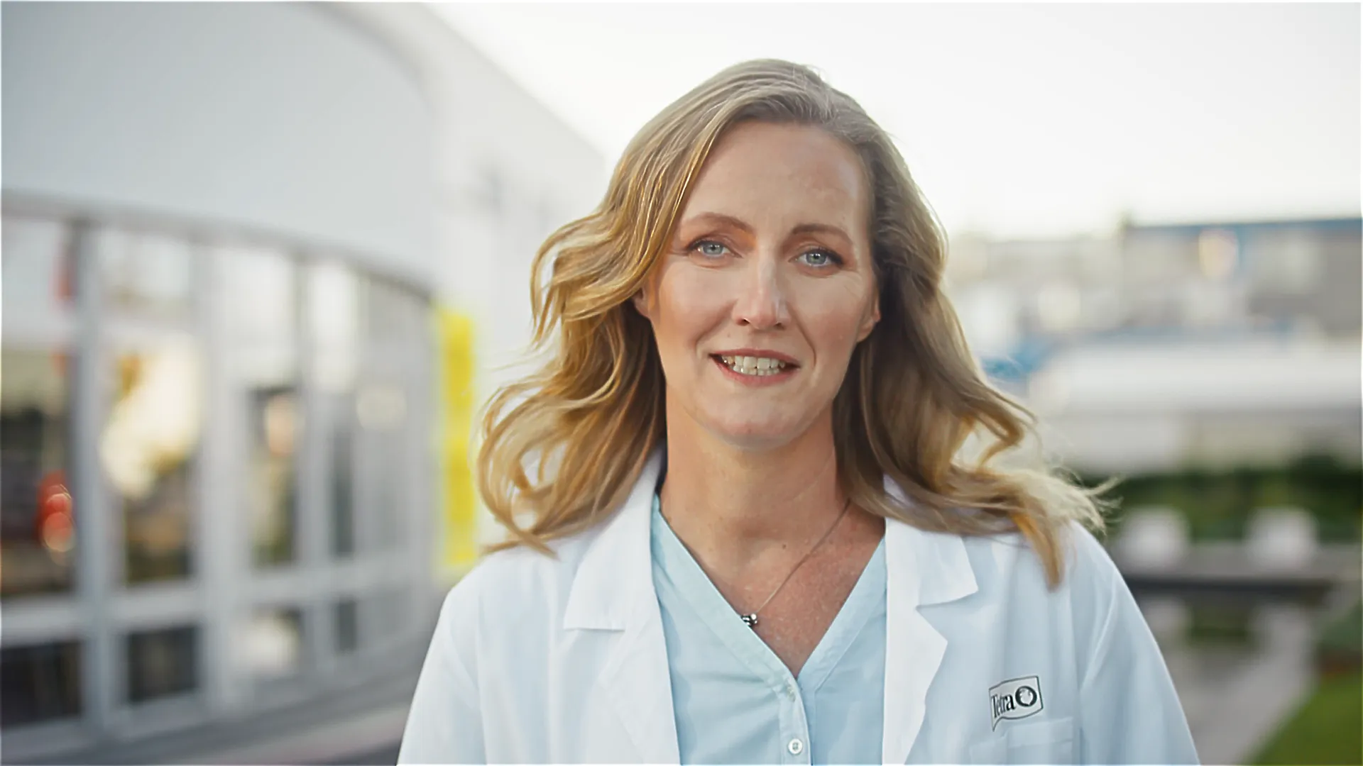 Porträt: Frau im weißen Laborkittel vor Firmengebäude, Expertin für Forschung und Nachhaltigkeit bei Tetra, lächelt in die Kamera.