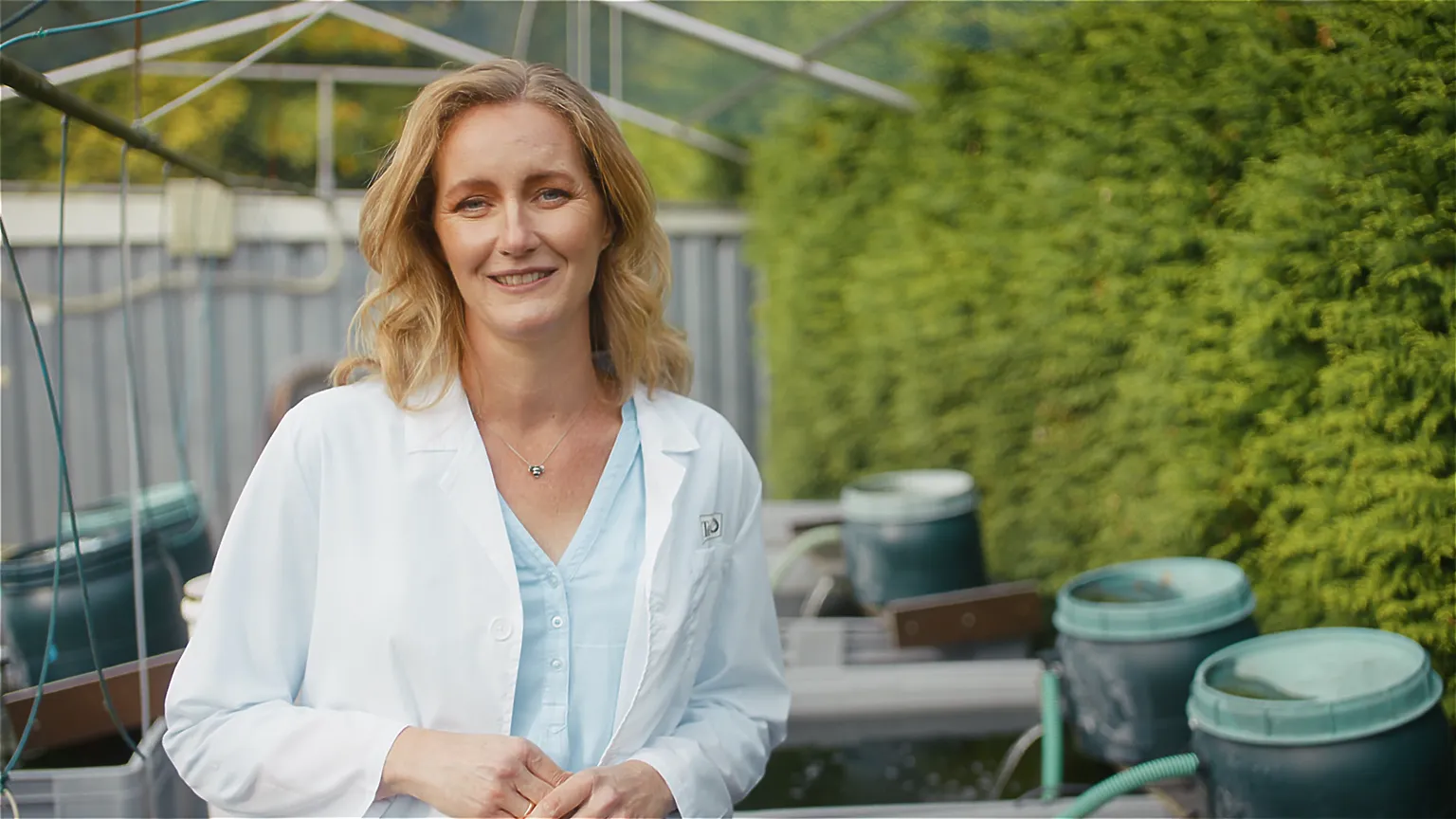Outdoor-Szene: Forscherin im weißen Kittel bei Testanlage im Freien mit Wasserbecken – Nachhaltigkeit und Aquakultur-Forschung.