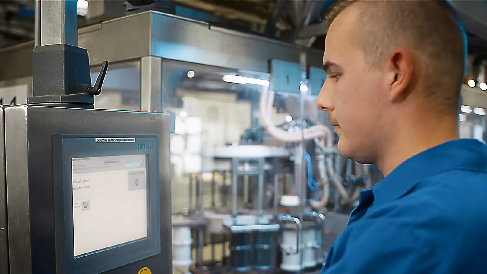 Produktionshalle: Mitarbeiter in blauem Kittel bedient Steuerung an einer digitalen Maschine für Fischfutter-Produktion.