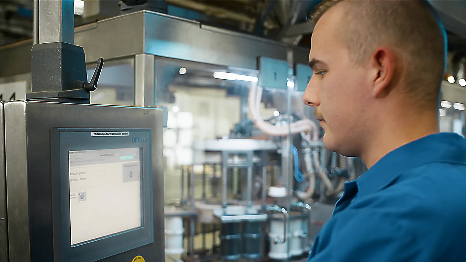 Produktionshalle: Mitarbeiter in blauem Kittel bedient Steuerung an einer digitalen Maschine für Fischfutter-Produktion.