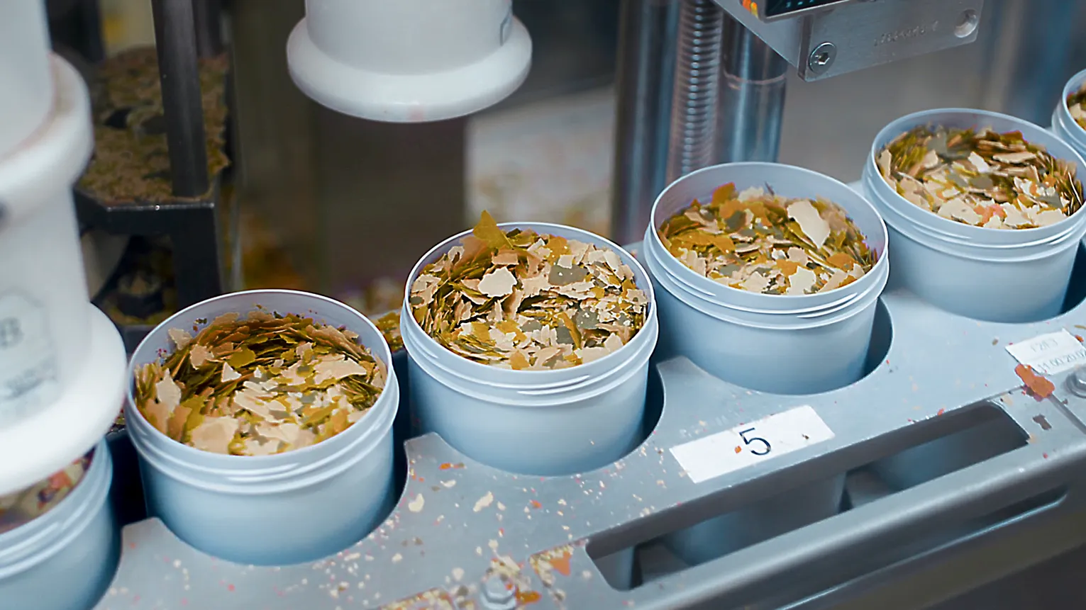 Produktionsaufnahme: Kunststoffdosen mit bunten Fischfutterflocken werden automatisch befüllt – nachhaltige Herstellung bei Tetra.