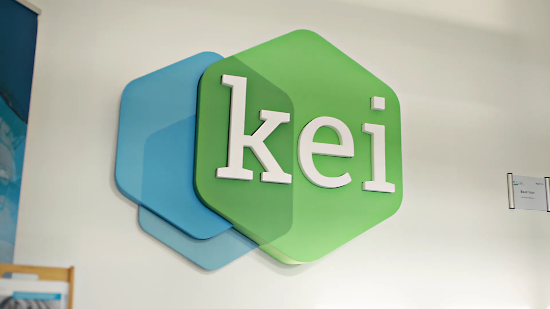 Großes KEI Logo in grüner und blauer Farbgestaltung an einer hellen Wand im Bürogebäude. Das moderne Design symbolisiert das Kompetenzzentrum Klimaschutz in der Industrie und die Förderung nachhaltiger Produktion.