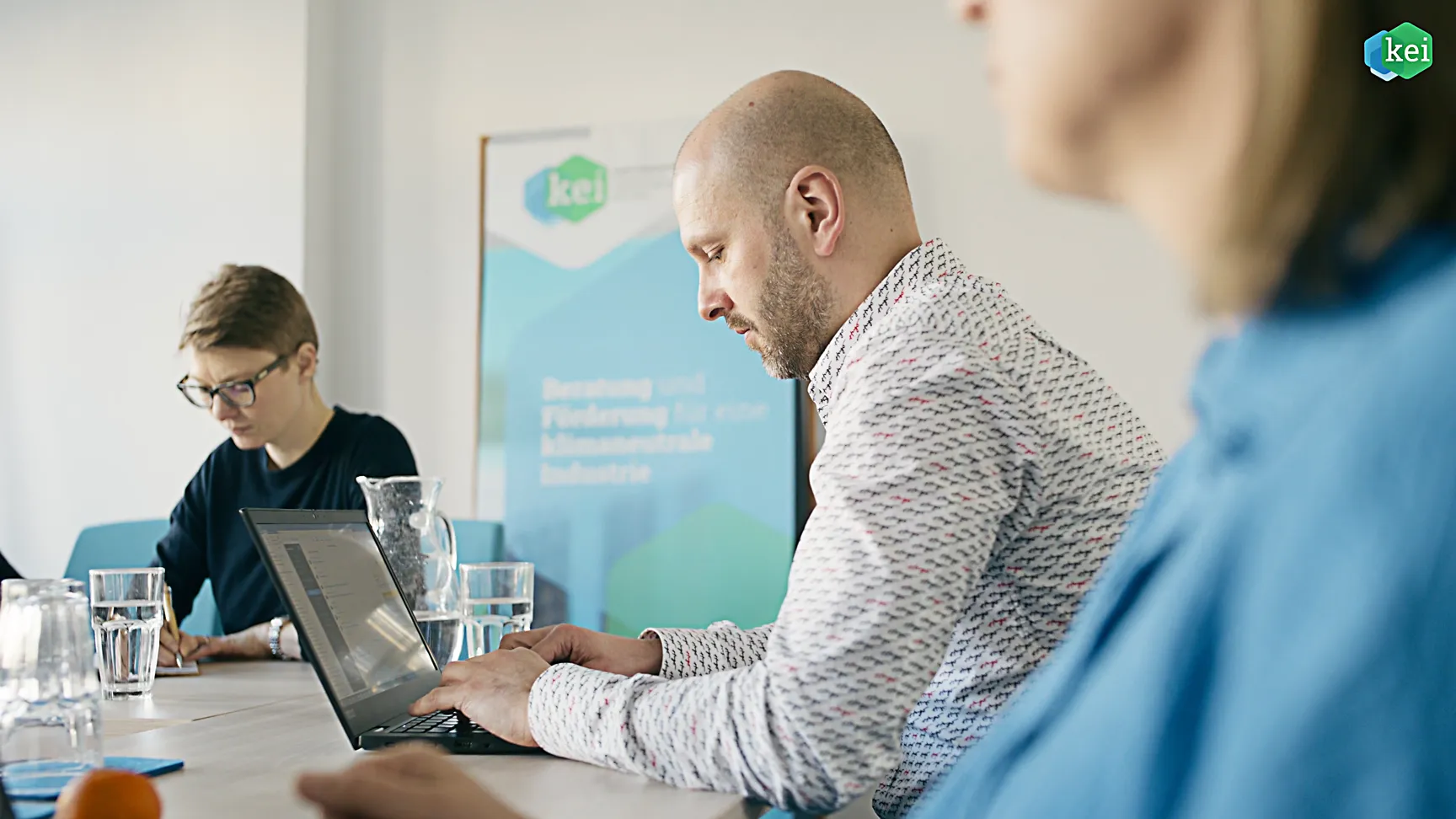 Mehrere Personen sitzen im Konferenzraum, arbeiten an Laptops und schreiben Notizen. Auf dem Tisch stehen Wassergläser und eine Karaffe. Im Hintergrund ein KEI-Banner mit dem Schriftzug Beratung und Förderung für klimaneutrale Industrie.