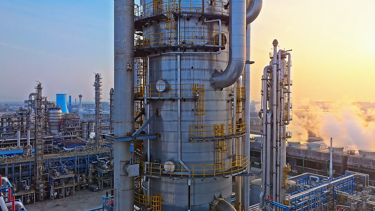 Industrieraffinerie mit großen Türmen, Rohrleitungen und Schornsteinen, aus denen Rauch aufsteigt. Die Szene wurde bei Sonnenaufgang aufgenommen und verdeutlicht die Relevanz von Klimaschutz, Dekarbonisierung und nachhaltiger Industrieproduktion.