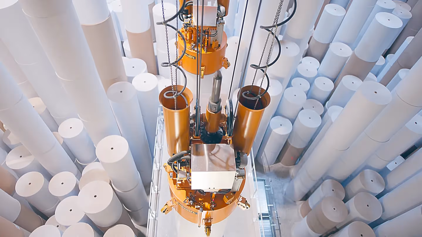 Ein automatisiertes Greifsystem hebt große Papierrollen in einer Fabrikhalle an. Im Hintergrund stapeln sich zahlreiche weiße Rollen in unterschiedlichen Höhen. Die Szene symbolisiert moderne Industrieprozesse und Effizienz in der Produktion.