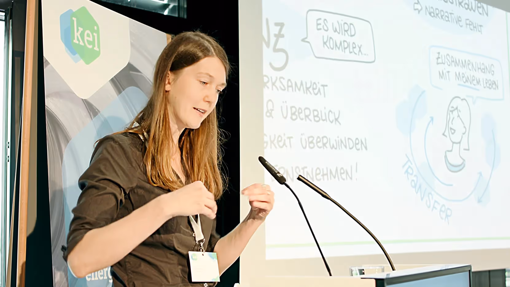 Junge Frau mit langen Haaren spricht an einem Rednerpult mit Mikrofon. Im Hintergrund eine Präsentationsfolie mit Text und Illustration sowie ein KEI-Banner mit Logo. Die Aufnahme zeigt Austausch, Wissenstransfer und klimafreundliche Technologien.