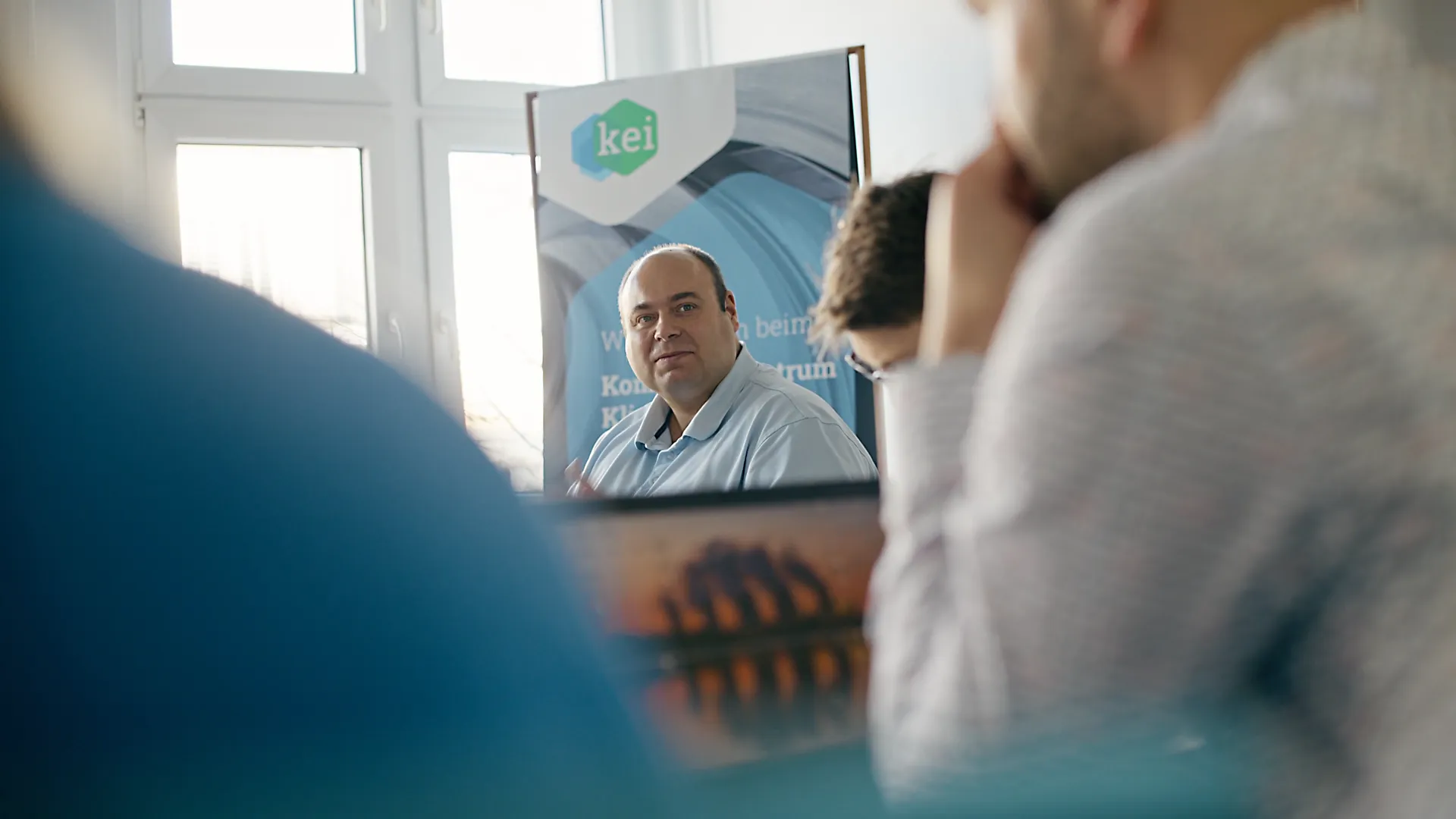Diskussion in einem Konferenzraum mit mehreren Teilnehmern. Im Vordergrund unscharfe Personen, im Hintergrund ein Mann mittleren Alters im blauen Hemd vor einem KEI-Banner. Die Szene zeigt Beratung, Förderung und Austausch für klimaneutrale Industrie.