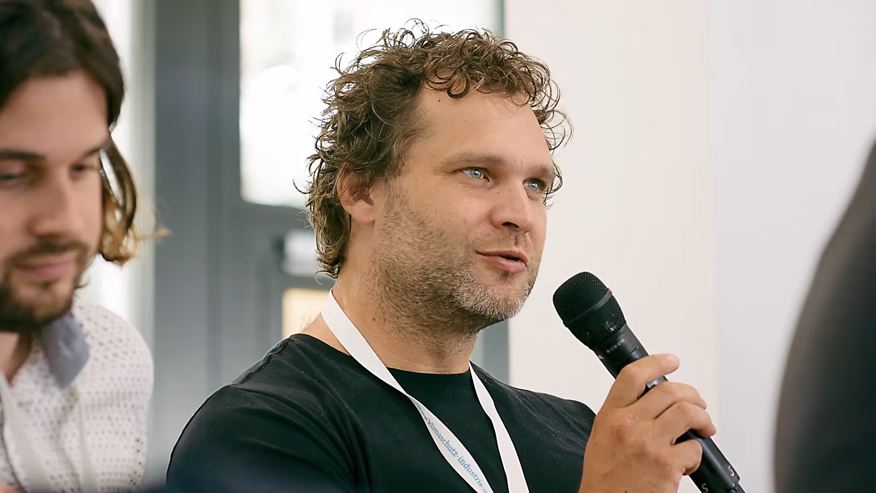 Mann mit lockigem Haar und schwarzem T-Shirt spricht in ein Mikrofon während einer Veranstaltung. Er trägt ein weißes Lanyard mit Namensschild und sitzt in einer Diskussionsrunde neben weiteren Teilnehmern zum Thema Nachhaltigkeit in der Industrie.