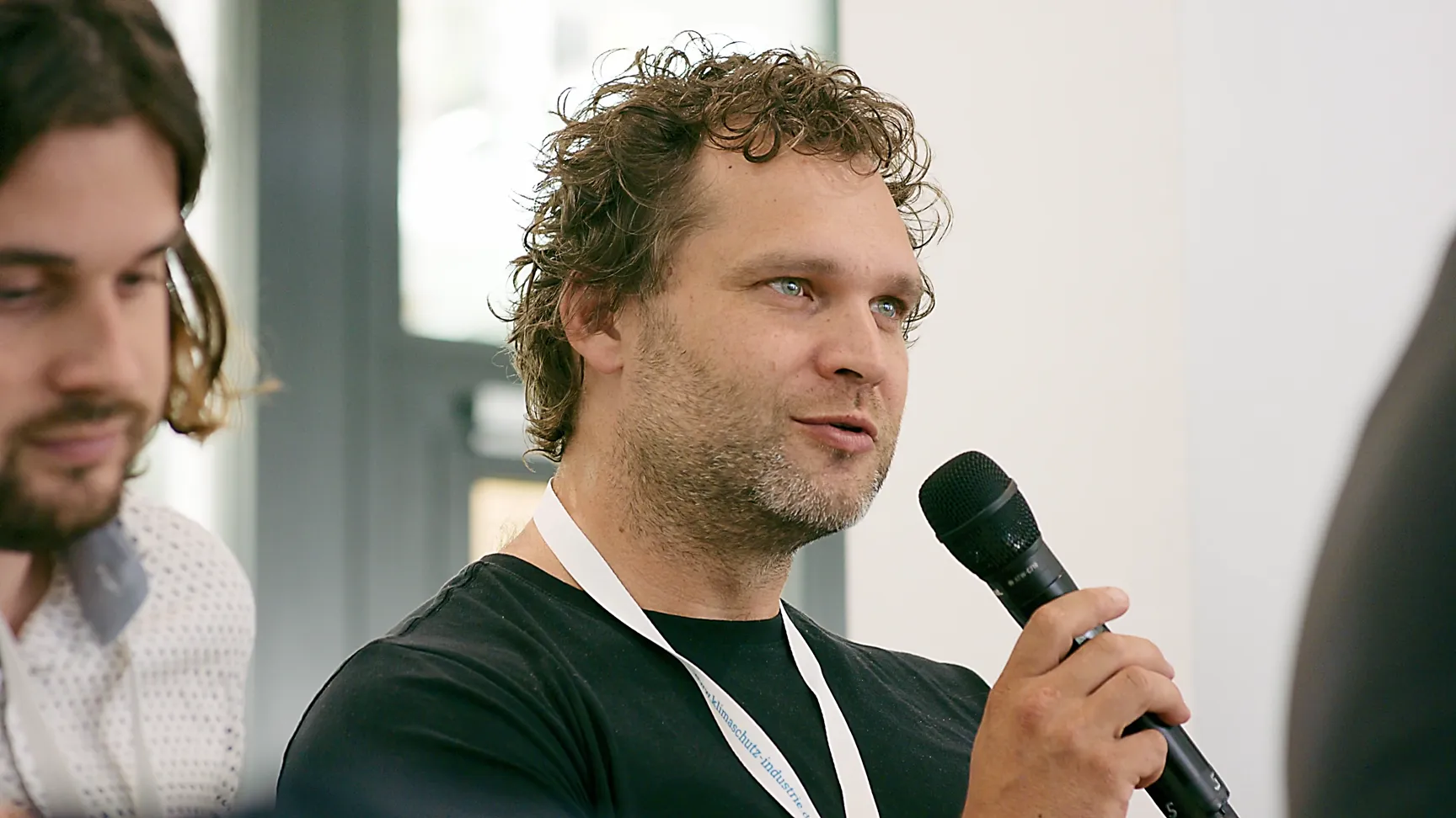 Mann mit lockigem Haar und schwarzem T-Shirt spricht in ein Mikrofon während einer Veranstaltung. Er trägt ein weißes Lanyard mit Namensschild und sitzt in einer Diskussionsrunde neben weiteren Teilnehmern zum Thema Nachhaltigkeit in der Industrie.