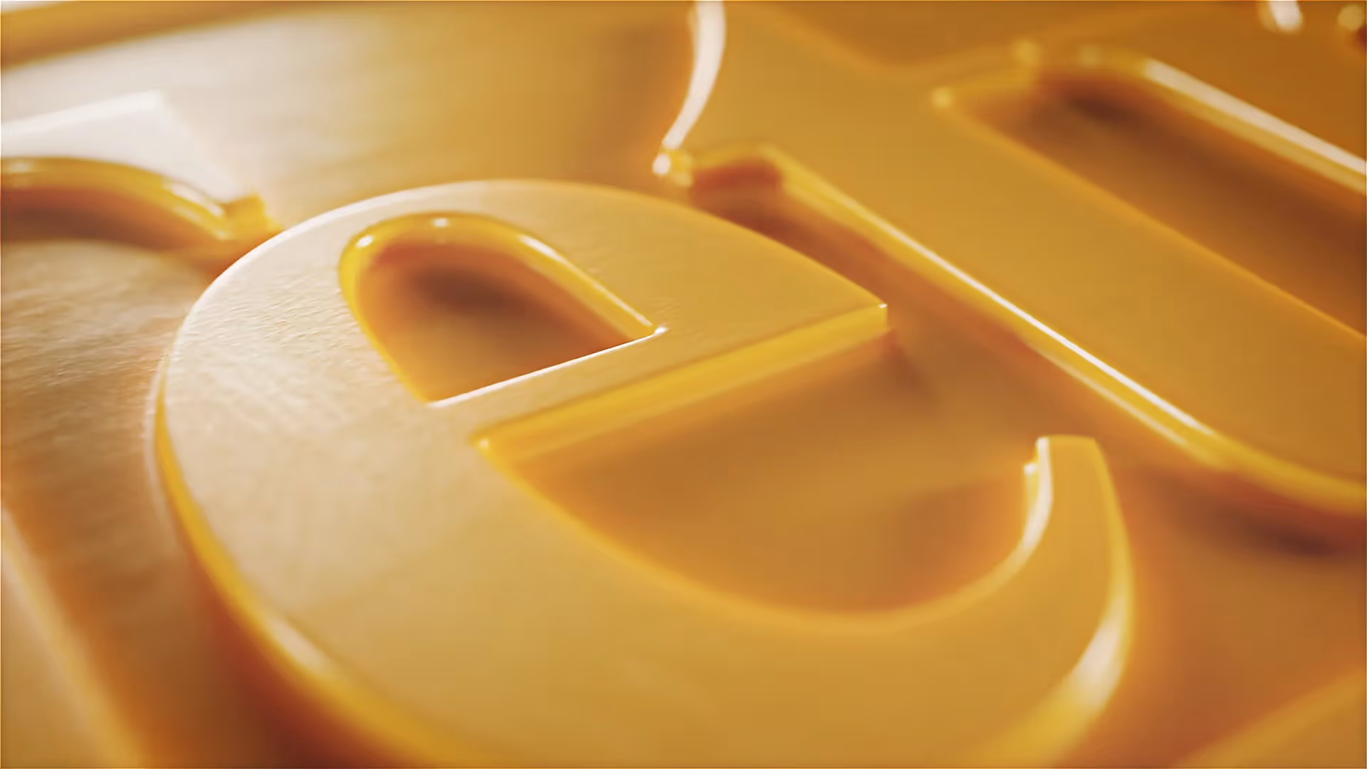 Makroaufnahme einer goldgelben 3D-Prägung: glänzender Buchstabe „e“ aus dem Logo von Tetra, detailreich beleuchtet mit weichen Lichtreflexen – moderne Produktanimation.