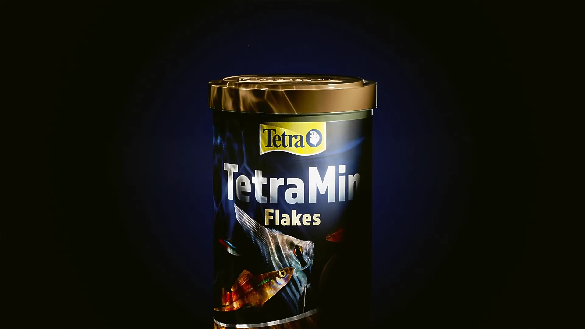 3D-Visualisierung einer TetraMin Flakes Dose mit Fischen auf blauem Etikett, goldener Deckel, freigestellt auf dunklem Hintergrund.