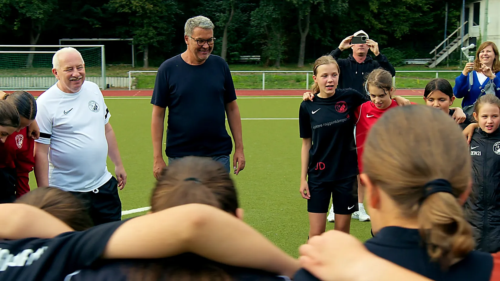 Kinder und Trainer stehen Arm in Arm im Kreis auf einem Fußballplatz, lachend und motiviert.