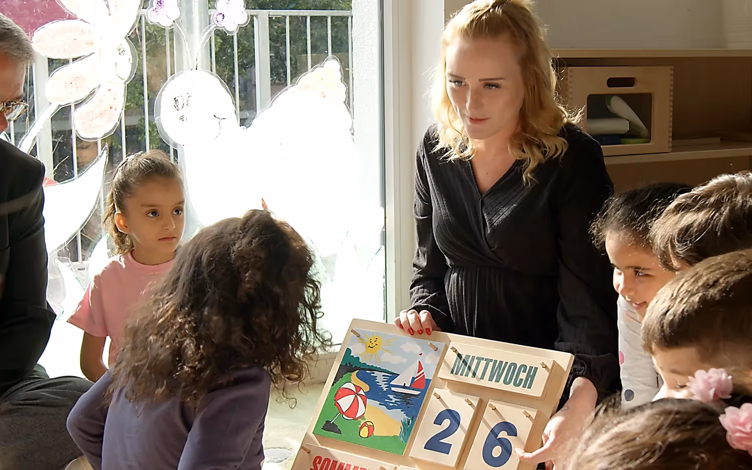 Eine Erzieherin zeigt Kindern in einer Kita einen Kalender mit Wochentagen und bunten Bildern.