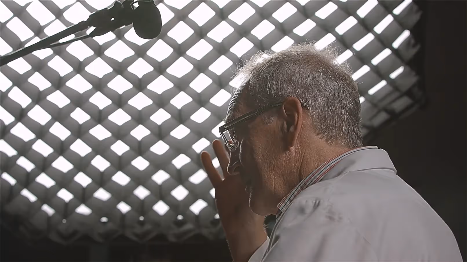 Interview-Szene: älterer Wissenschaftler im weißen Kittel mit Brille im Studio, Mikrofon über Kopf, Lichtgitter im Hintergrund – Expertenaussage für Forschung und Entwicklung.