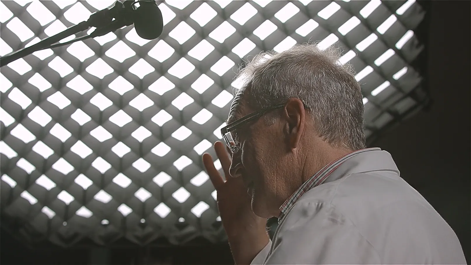 Interview-Szene: älterer Wissenschaftler im weißen Kittel mit Brille im Studio, Mikrofon über Kopf, Lichtgitter im Hintergrund – Expertenaussage für Forschung und Entwicklung.