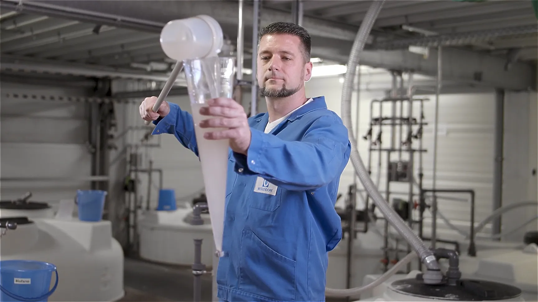 Laboraufnahme: Mann im blauen Kittel misst Flüssigkeit mit großem Trichter-Messzylinder, industrielle Forschungsumgebung mit Rohren und Tanks – Wasser- und Produktanalyse.