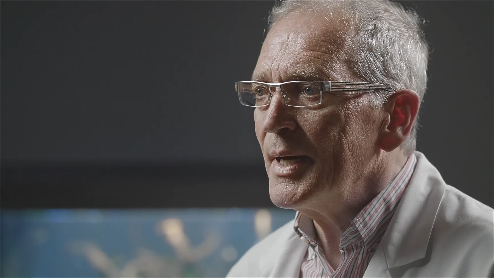 Interviewaufnahme: Wissenschaftler im weißen Laborkittel vor dunklem Hintergrund spricht über Forschung, Brille und graue Haare, Nahaufnahme für Dokumentation.