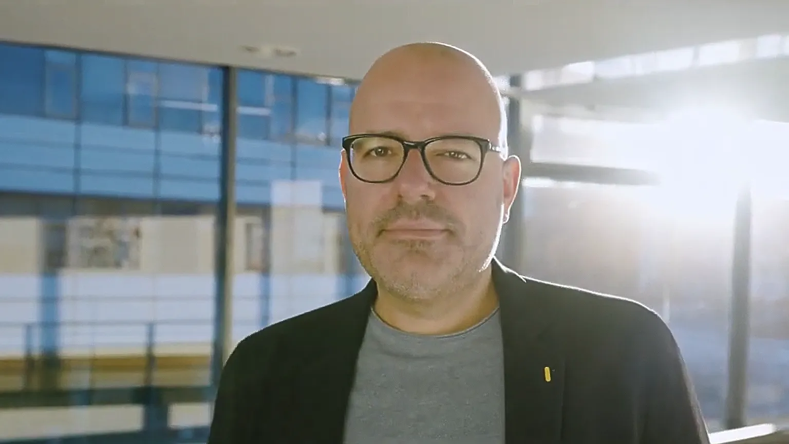 Porträt eines Mannes mit Brille im hellen Laborgebäude, Sonnenlicht fällt durch die Fenster.