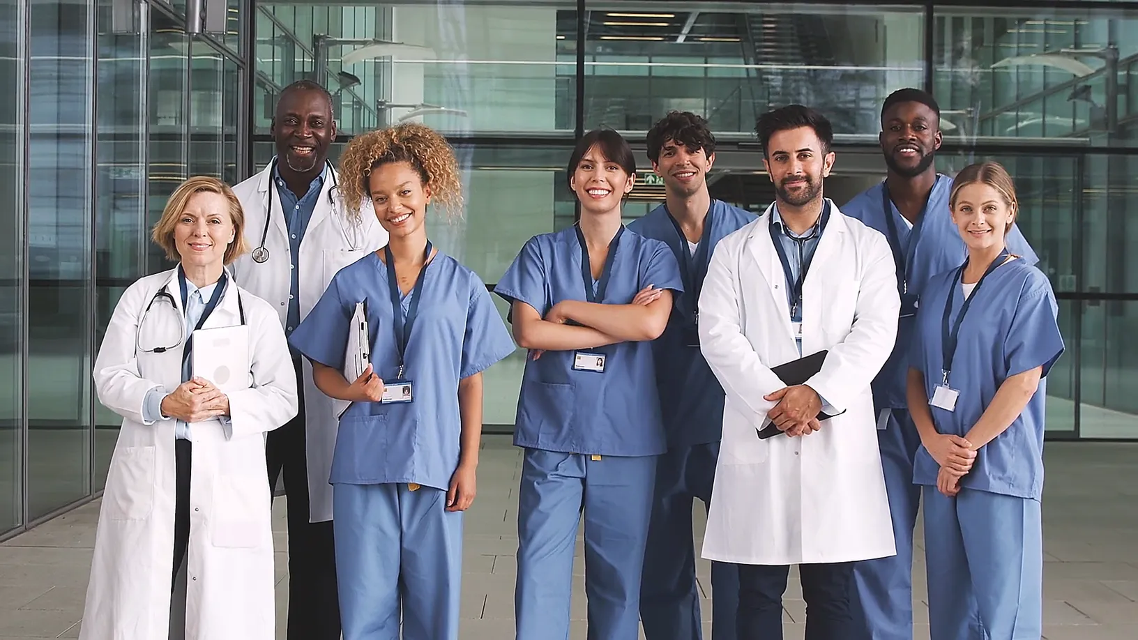 Vielfältiges Team aus sieben medizinischen Fachkräften in Kitteln und OP-Bekleidung lächelt vor einem modernen Klinikgebäude.
