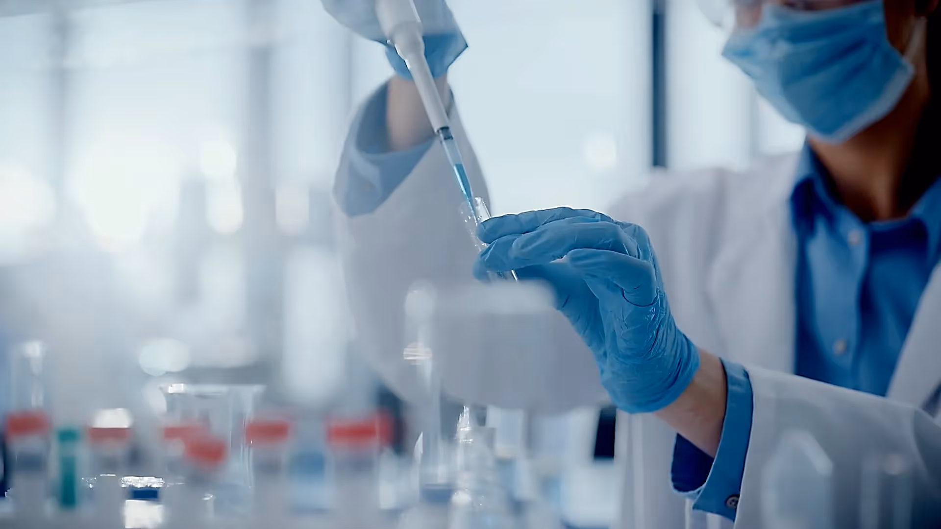 Wissenschaftler im Labor mit Maske und Handschuhen pipettiert blaue Flüssigkeit in Reagenzglas.