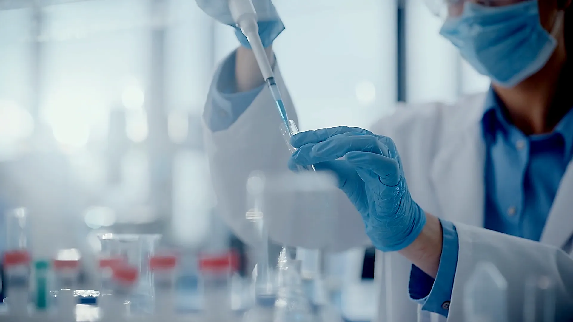 Wissenschaftler im Labor mit Maske und Handschuhen pipettiert blaue Flüssigkeit in Reagenzglas.