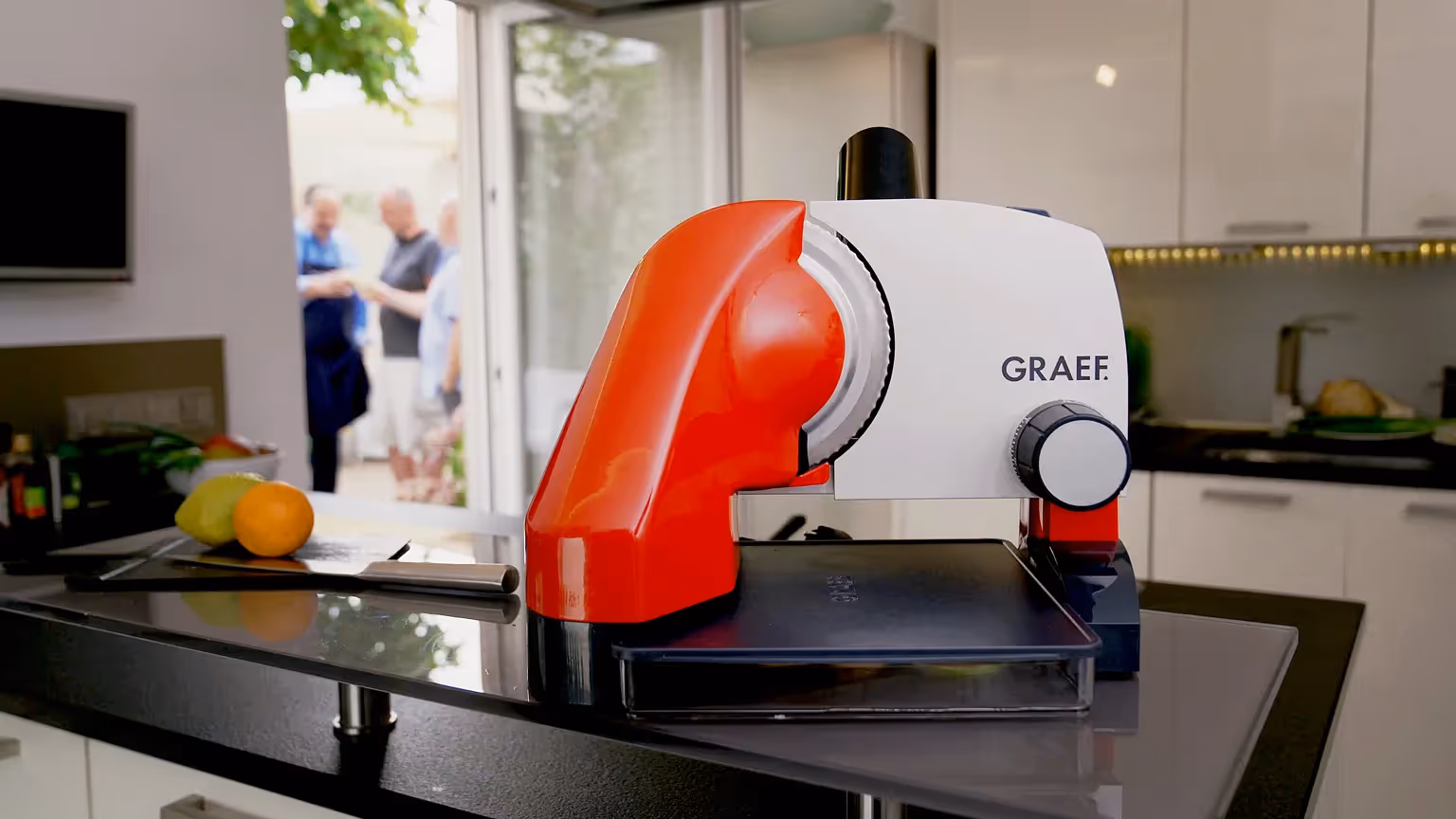 Graef Allesschneider in Rot-Weiß steht auf einer modernen Küchenarbeitsplatte; im Hintergrund offene Terrassentür mit unscharfen Personen, die sich unterhalten.