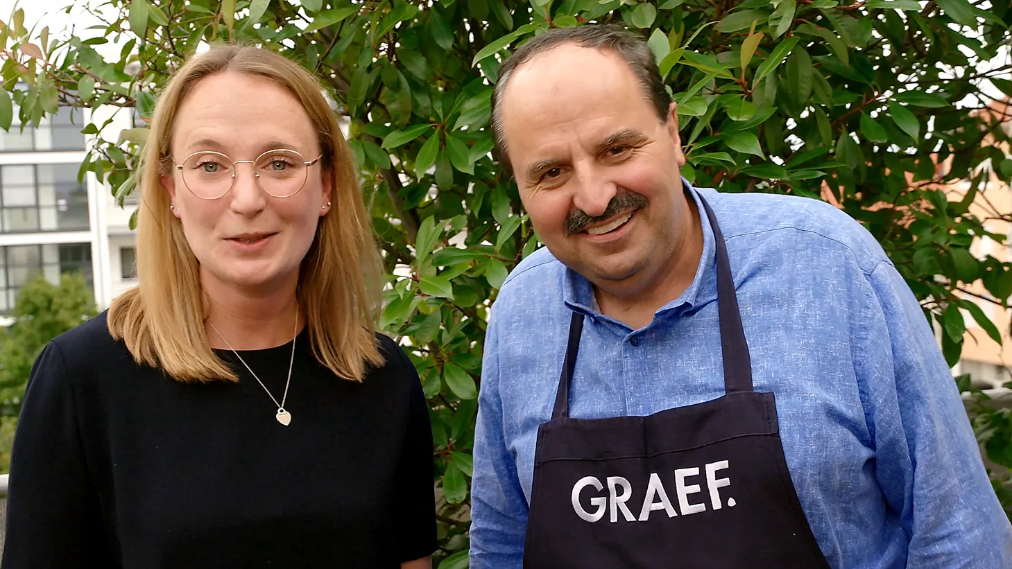 Zwei Personen stehen nebeneinander im Freien; links eine Frau mit Brille und schwarzem Oberteil, rechts ein Mann im blauen Hemd mit schwarzer Schürze mit weißem „GRAEF“-Logo, beide lächeln in die Kamera.