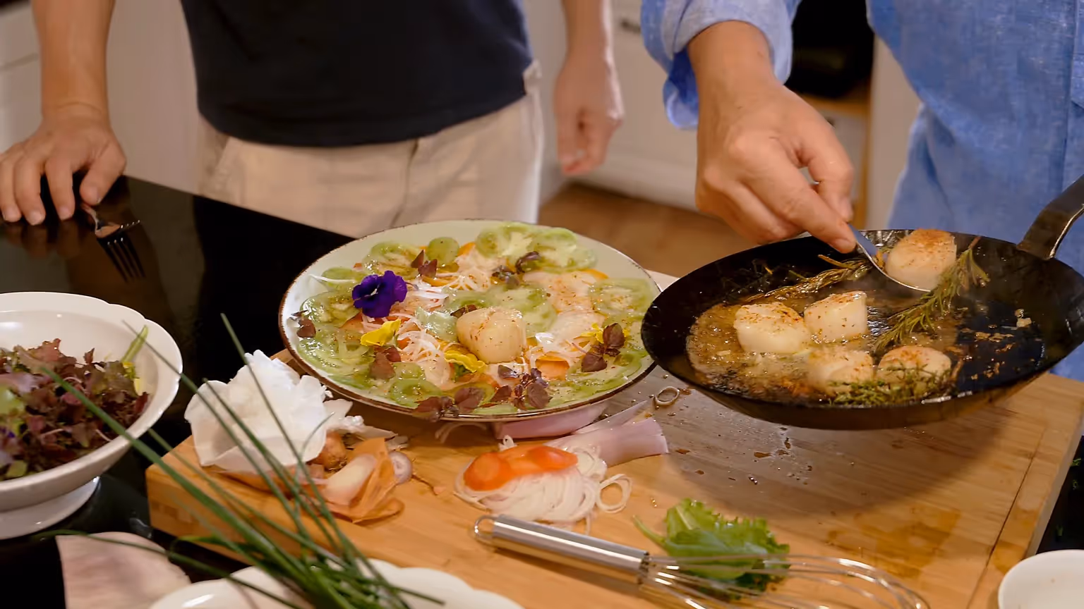 Ein Teller mit dekorativ angerichteten Jakobsmuscheln, Gurkenscheiben und essbaren Blüten; daneben eine Pfanne mit gebratenen Jakobsmuscheln, die mit Kräutern in Butter schäumen.
