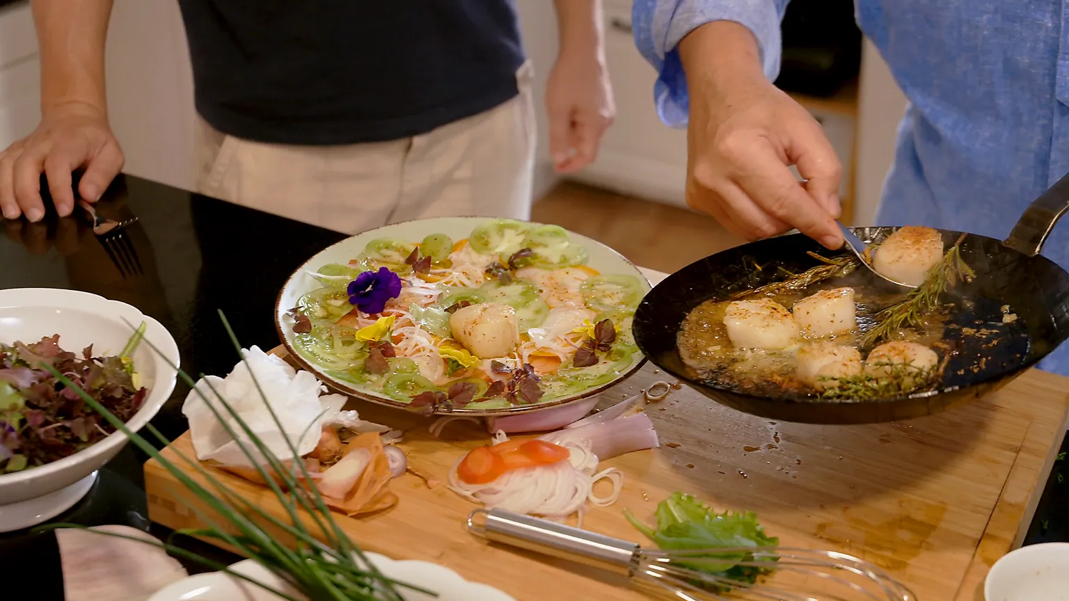 Ein Teller mit dekorativ angerichteten Jakobsmuscheln, Gurkenscheiben und essbaren Blüten; daneben eine Pfanne mit gebratenen Jakobsmuscheln, die mit Kräutern in Butter schäumen.