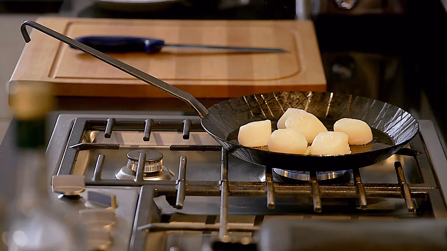 Eine schwarze Pfanne mit mehreren Jakobsmuscheln brät auf einem Gasherd; im Hintergrund ein Schneidebrett mit Messer.