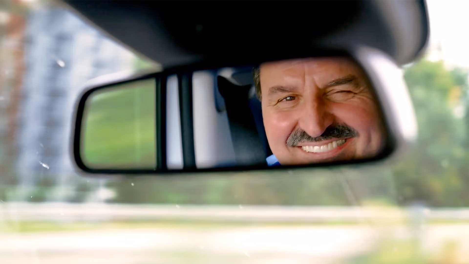 Ein Mann mit Schnurrbart lächelt fröhlich und zwinkert in den Rückspiegel eines Autos – ein humorvoller, charmanter Moment auf der Fahrt.