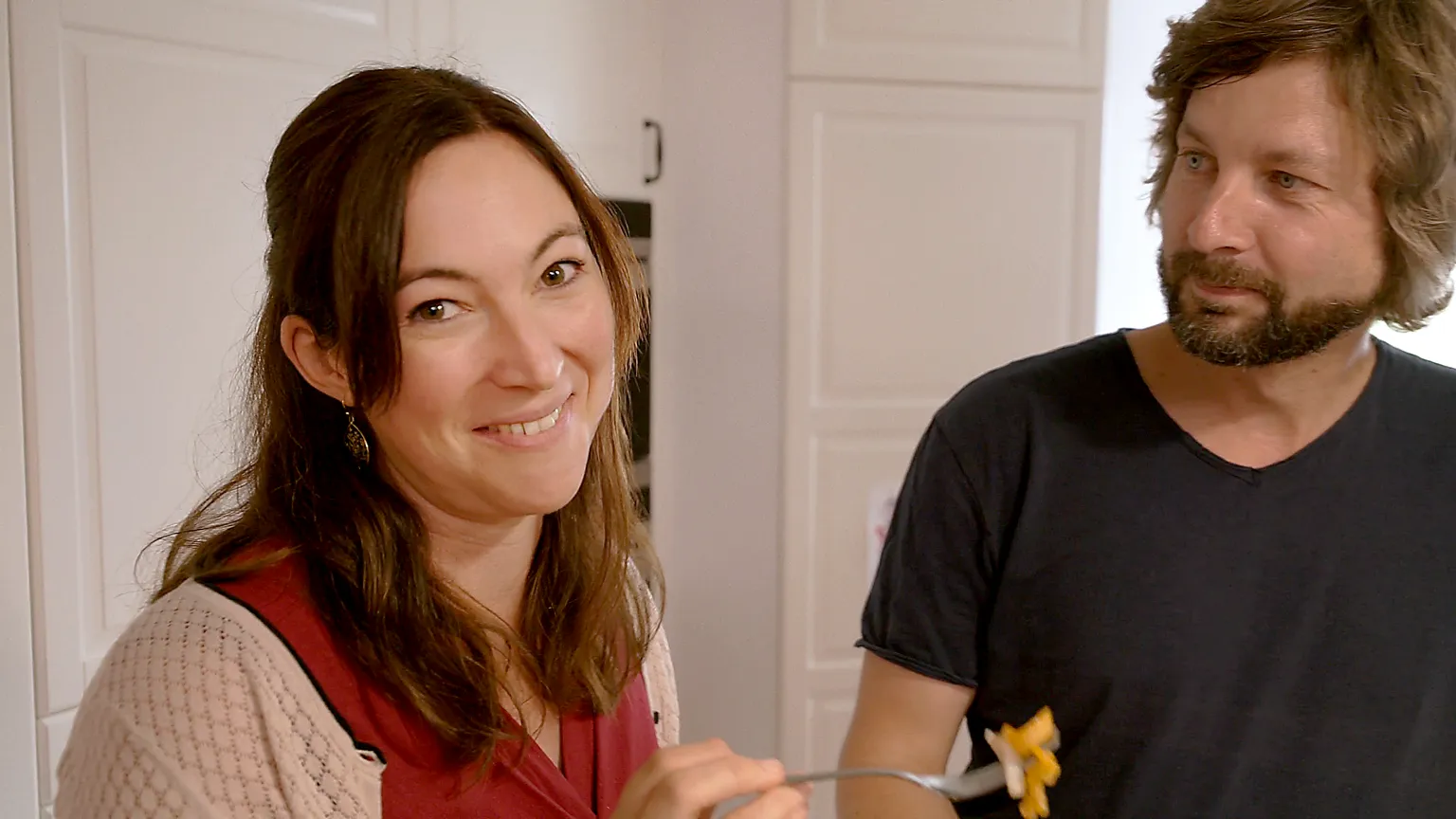 Eine Frau lächelt in die Kamera und hält eine Gabel mit Essen, während ein Mann sie daneben aufmerksam ansieht – eine Szene voller Genuss und Geselligkeit.