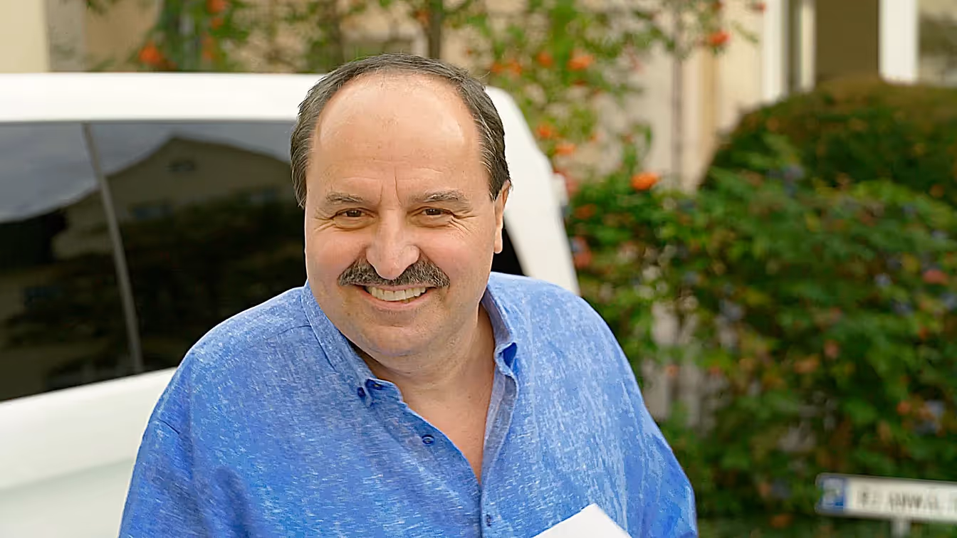 Ein Mann im blauen Hemd steht lachend vor einem weißen Fahrzeug, der Blick freundlich und offen – eine einladende Szene im Freien.